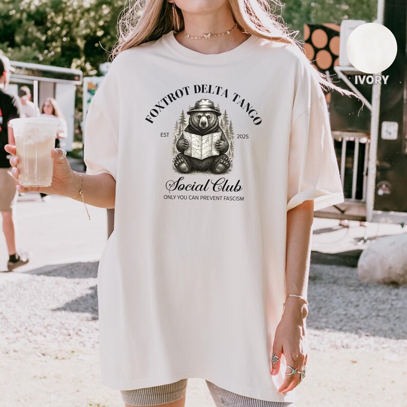 Puede incluir: Una camiseta blanca de gran tama&ntilde;o con un gr&aacute;fico de un oso con un sombrero de guardabosques y leyendo un mapa. El gr&aacute;fico est&aacute; rodeado de texto que dice "Fox Trot Delta Tango Est 2025 Social Club Only You Can Prevent Fascism".