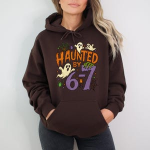 May include: A dark brown hooded sweatshirt with the phrase "Haunted by 6-7" in orange, purple, and green. The design includes ghost and spiderweb graphics, perfect for Halloween.