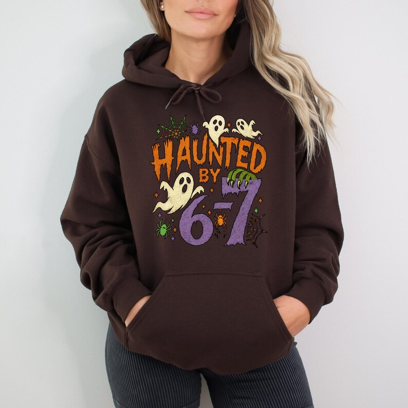 May include: A dark brown hooded sweatshirt with the phrase "Haunted by 6-7" in orange, purple, and green. The design includes ghost and spiderweb graphics, perfect for Halloween.