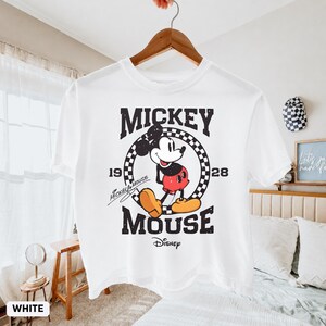 May include: White t-shirt featuring a vintage Mickey Mouse graphic. The design includes the words "Mickey Mouse" and "Disney" with the years "19" and "28". The shirt is hanging on a wooden hanger.