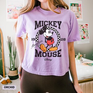 May include: Orchid-colored t-shirt featuring a graphic of Mickey Mouse. The design includes the words "Mickey Mouse" and "Disney", along with the years "1928" and "19". The shirt has short sleeves.