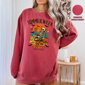 May include: Crimson sweatshirt with a graphic featuring a skeleton relaxing in a beach chair with a cocktail, pumpkins, and the words "Summerween Vibes." The design includes bats, a sunset, and the phrase "Forever Spooky."
