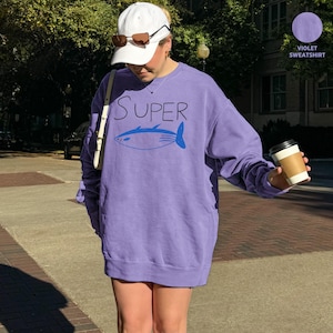May include: A violet sweatshirt featuring the word "SUPER" in black and a blue fish design. The long-sleeved sweatshirt is a solid colour. The model is wearing a white cap and sunglasses.