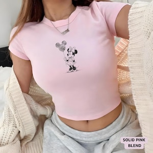 May include: A light pink crop top featuring a black and white illustration of Minnie Mouse holding a balloon. The shirt has short sleeves and a crew neck. The text "SOLID PINK BLEND" is in a white box at the bottom right.