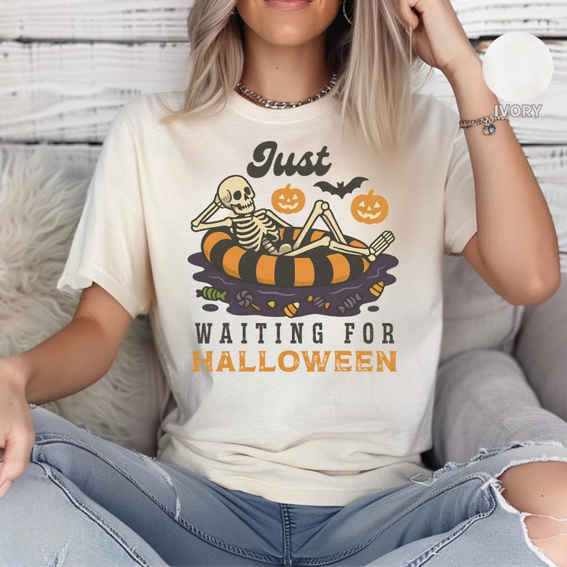 May include: Cream-colored t-shirt with a Halloween-themed graphic. The design features a skeleton relaxing on an orange and black inner tube, with pumpkins and a bat. The text reads "Just Waiting for Halloween."