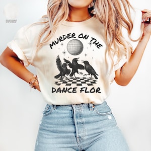 May include: Ivory-colored t-shirt featuring the text "MURDER ON THE DANCE FLOR" with a disco ball and three black crow illustrations. The shirt has a crew neck and short sleeves. The shirt is made of a soft, comfortable material.