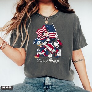 May include: Dark gray t-shirt featuring a patriotic design with Mickey Mouse, Donald Duck, and Mickey Mouse holding a flag. The shirt has the text "250 Years" in white. The design incorporates red, white, and blue colors.