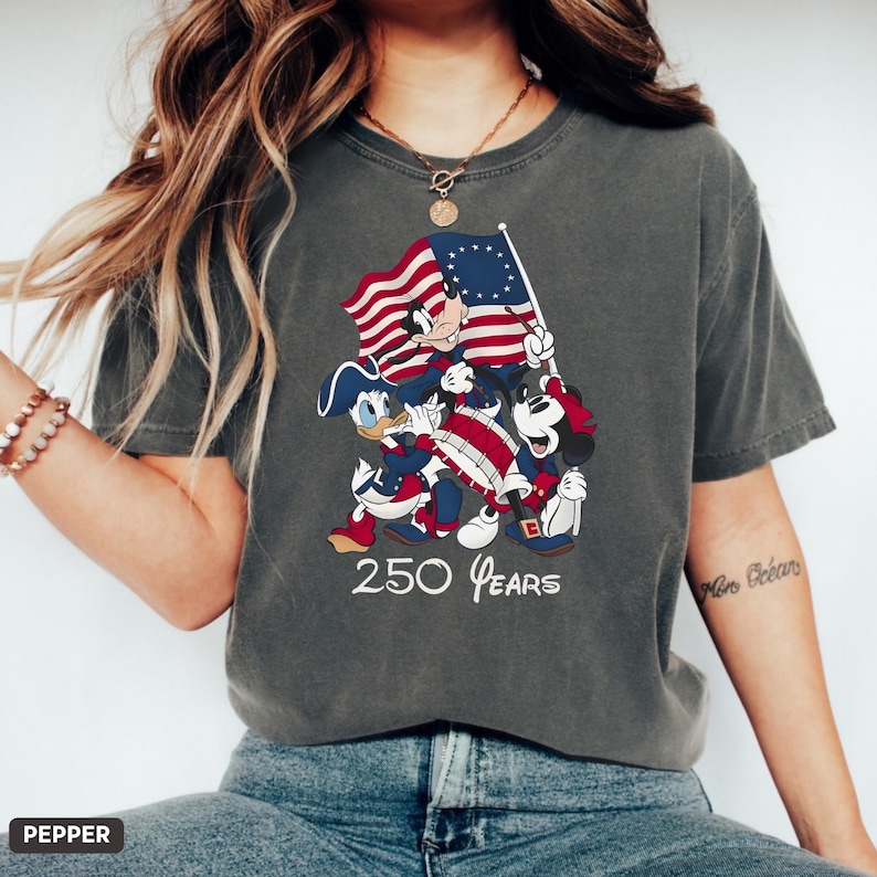 May include: Dark gray t-shirt featuring a patriotic design with Mickey Mouse, Donald Duck, and Mickey Mouse holding a flag. The shirt has the text "250 Years" in white. The design incorporates red, white, and blue colors.