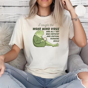May include: A white t-shirt featuring a green frog cartoon holding a cup of coffee. The text on the shirt reads "I caught the woke mind virus and all I got was empathy and critical thinking skills."