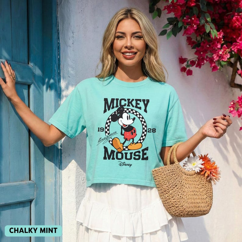 May include: A mint green crop top featuring a Mickey Mouse graphic. The graphic includes the words "Mickey Mouse" and the years "1928" and "1928". The model is also wearing a white skirt and carrying a woven handbag with flowers.