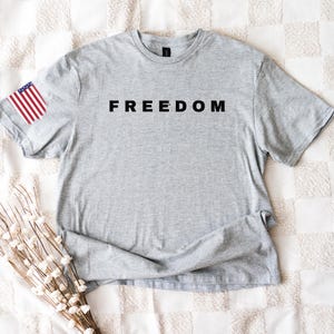 K&ouml;nnte beinhalten: Ein meliertes graues T-Shirt mit dem Wort "FREEDOM" in Schwarz auf der Brust aufgedruckt. Auf dem linken &Auml;rmel befindet sich ein kleines amerikanisches Flaggen-Patch. Das Shirt besteht aus weichem Material und liegt flach auf einer wei&szlig;en, gemusterten Oberfl&auml;che.