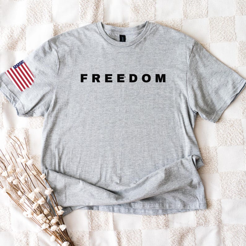 K&ouml;nnte beinhalten: Ein meliertes graues T-Shirt mit dem Wort "FREEDOM" in Schwarz auf der Brust aufgedruckt. Auf dem linken &Auml;rmel befindet sich ein kleines amerikanisches Flaggen-Patch. Das Shirt besteht aus weichem Material und liegt flach auf einer wei&szlig;en, gemusterten Oberfl&auml;che.