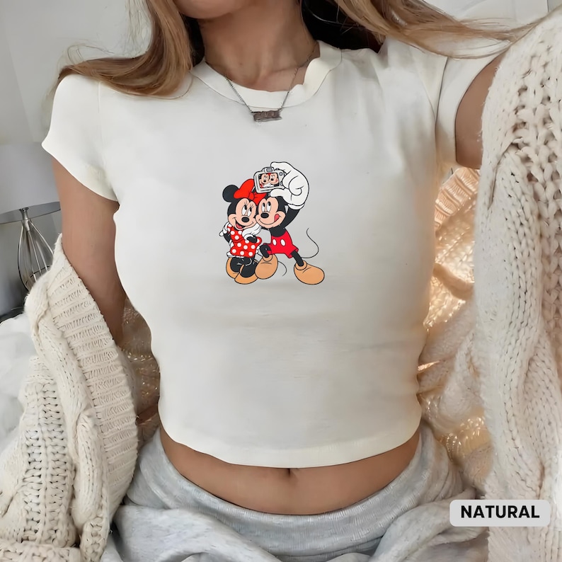 May include: Cream-colored, short-sleeved crop top featuring a graphic of Mickey and Minnie Mouse. Mickey is holding a camera, and Minnie is smiling. The top has a rounded neckline and is made of a soft material. The word "NATURAL" is at the bottom right.
