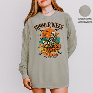 May include: Sandstone long-sleeve shirt with a graphic featuring a skeleton relaxing in a beach chair with a cocktail, pumpkins, and the words "Summerween Vibes Forever Spooky". The design is in orange, black, and yellow.