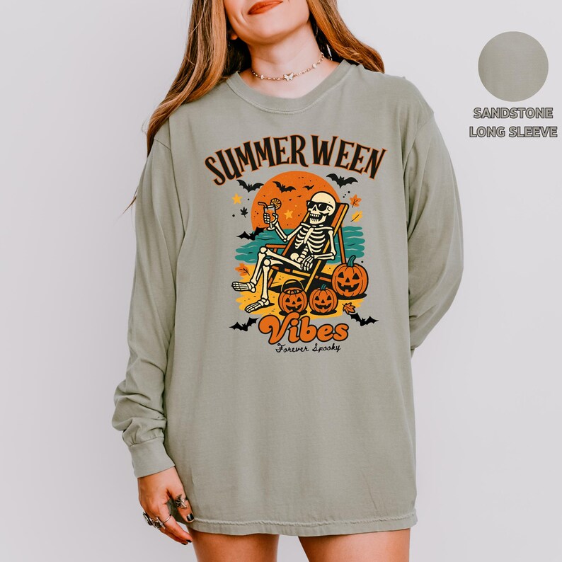May include: Sandstone long-sleeve shirt with a graphic featuring a skeleton relaxing in a beach chair with a cocktail, pumpkins, and the words "Summerween Vibes Forever Spooky". The design is in orange, black, and yellow.