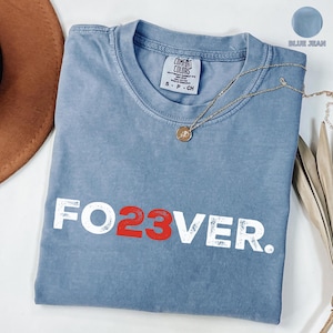 May include: Blue jean-colored t-shirt with the text "FO23VER" in white and red. The shirt has a crew neck and short sleeves. A gold necklace with a pendant is draped across the shirt.