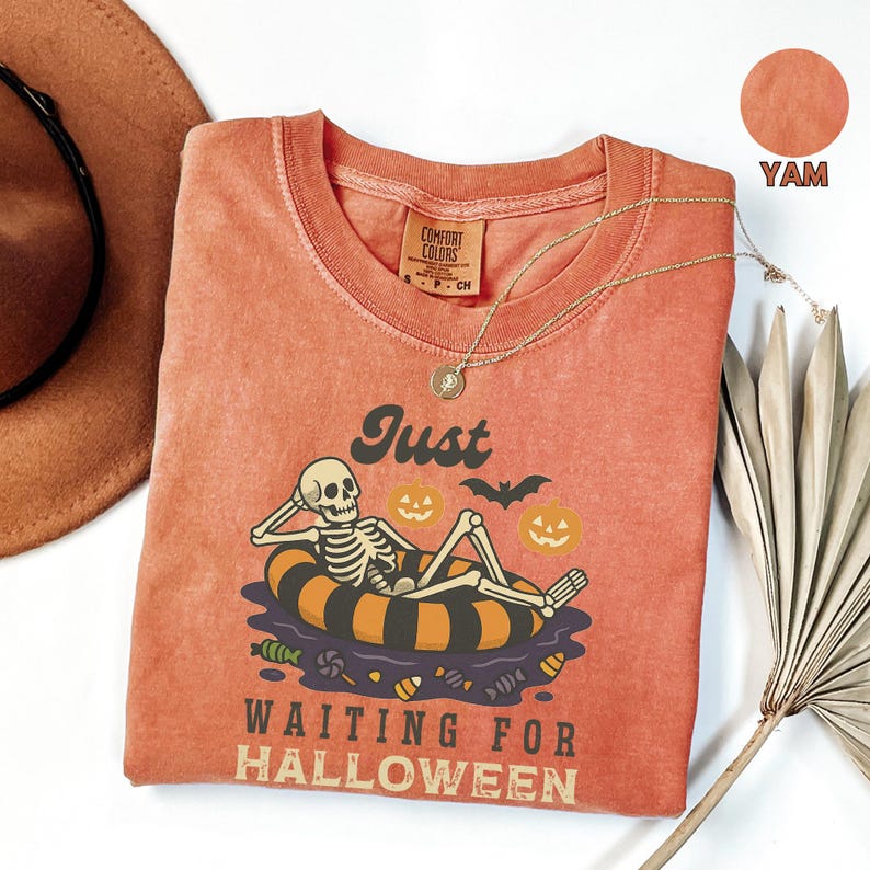 May include: A yam-colored, crew neck t-shirt with a Halloween-themed graphic. The graphic features a skeleton relaxing on an inner tube, surrounded by pumpkins, candy, and a bat. The text reads "Just Waiting for Halloween."
