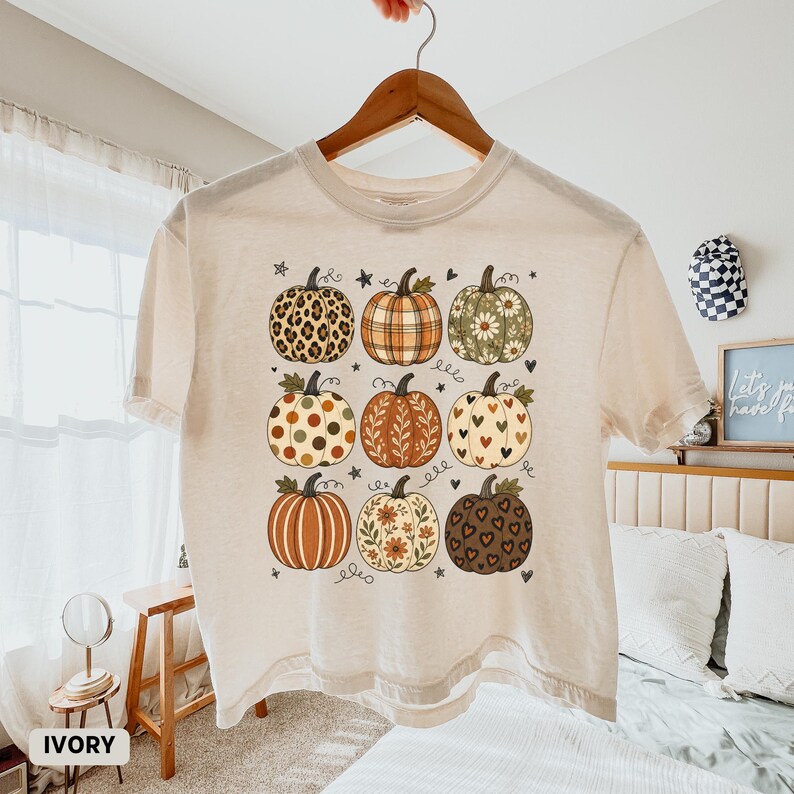May include: An ivory short-sleeve t-shirt featuring a graphic print of nine pumpkins in a grid layout. Each pumpkin displays a unique pattern, including leopard print, plaid, floral, polka dots, and hearts, accented by small stars and hearts.
