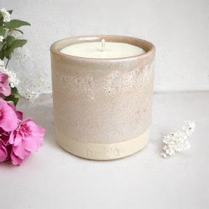 May include: A white ceramic candle jar with a light pink glaze and a white wax candle inside. The jar has the word "nudia" embossed on the bottom.