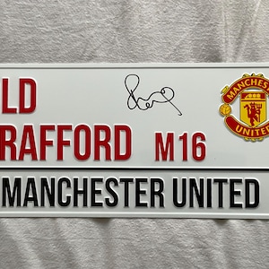 Könnte beinhalten: Weißes rechteckiges Schild mit roter und schwarzer Schrift. Auf dem Schild steht "OLD TRAFFORD M16 MANCHESTER UNITED". Enthält eine Unterschrift und das Manchester United Logo.