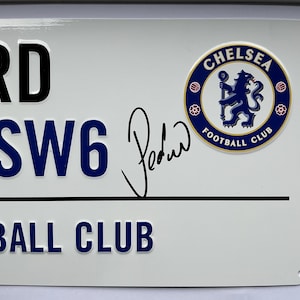 May include: White metal sign featuring the text "FORD", "SW6", and "FOOTBALL CLUB" in black and blue. The Chelsea Football Club logo is on the right. A signature is present. The sign has rounded corners and mounting holes.