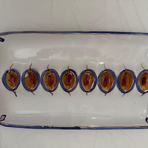 May include: A white rectangular ceramic serving platter with a blue and gold trim. The platter has a row of eight painted brown and red plums.