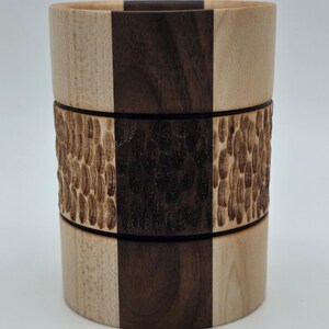 Könnte beinhalten: Ein Holzbecher mit einem einzigartigen Design. Der Becher hat abwechselnde vertikale Paneele aus hellem und dunklem Holz. Ein strukturiertes Band mit einem Muster umschließt die Mitte. Der Becher ist zylindrisch und steht aufrecht.
