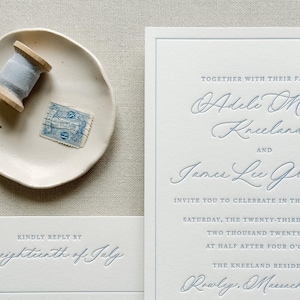 May include: Elegant wedding invitation suite with embossed blue text on white cardstock. Includes RSVP card, invitation, and details card. A small dish holds a spool of blue ribbon and a vintage postage stamp.