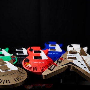 May include: A collection of six miniature guitar picks, each shaped like a different guitar model. The picks are made of plastic and feature different colors, including green, black, red, blue, and white. The picks are all labeled with the name of the guitar manufacturer, such as "G. Paul Rich" and "Dyer-Jork".