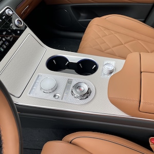 May include: The interior center console of a vehicle features tan quilted leather seats, a silver textured panel with two circular cup holders, and two metallic control dials. A digital touchscreen display shows climate settings at 65 degrees.