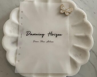 Puede incluir: Una hoja de papel blanca translúcida con una pegatina de mariposa dorada en la esquina superior derecha. El papel tiene agujeros en el lado izquierdo y el texto "Dawning Horizon" escrito en cursiva negra. Debajo del texto está la frase "Dream. Plan. Achieve." en cursiva negra más pequeña.