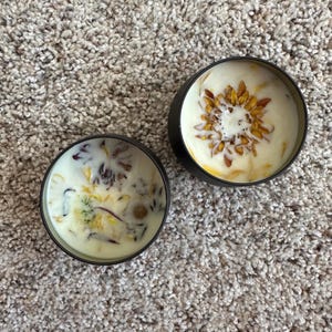 May include: Two black candle jars with white wax and dried flowers. The flowers are pressed into the wax and are visible on the surface. The candle jars are on a light brown carpet.