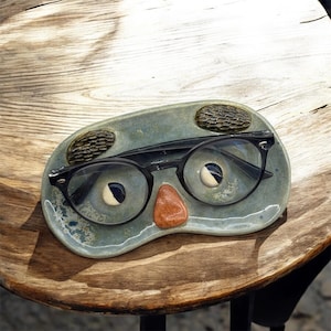 May include: A blue ceramic dish shaped like a face with glasses, a nose, and eyebrows. The dish is designed to hold glasses.