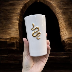 May include: A white ceramic tumbler with a gold snake design. The cylindrical cup is held in a hand, showcasing the embossed serpent. The background features a dark archway.