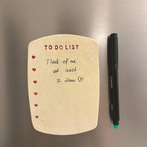 Puede incluir: Un imán rectangular beige con una lista de tareas, detalles de corazones rojos y el texto escrito a mano "Think of me at least 7 times ❤". Un rotulador negro con punta turquesa descansa junto al imán.