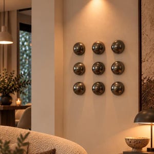 May include: A set of nine circular, metallic, dark-toned wall-mounted decorative spheres arranged in a three-by-three grid pattern on a light-coloured wall, illuminated by a single overhead cylindrical light fixture.