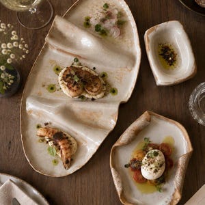 May include: A set of three off-white ceramic serving dishes with irregular, organic shapes and textured edges. The largest platter features two raised ridges, while the smaller rectangular bowl and the curved side plate hold prepared food items.