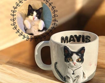 Puede incluir: Taza de cerámica blanca con un diseño de gato blanco y negro, el nombre "MAVIS" en letras negras en relieve y una pequeña huella de pata. Una imagen de un gato en un marco circular está en el fondo. La taza tiene un asa.