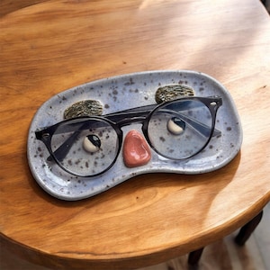 Può includere: Un portaocchiali in ceramica blu a forma di viso con un paio di occhiali con montatura nera in cima. Il viso ha un naso rosa e sopracciglia nere.