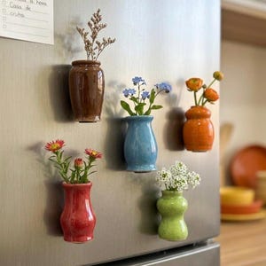 以下が含まれることがあります： 金属面に固定された5つの小さなカラフルな陶器製花瓶。それぞれに異なる花が生けられています。花瓶は茶色、青、オレンジ、赤、ライトグリーンの色で、滑らかで光沢のある仕上げと、花を飾るための様々な丸みを帯びた形状が特徴です。
