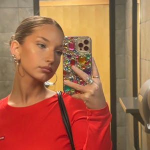 May include: A red long-sleeve top. The person is holding a bedazzled phone case with colorful gems. The person is wearing gold hoop earrings and rings. The background is a bathroom with a mirror.