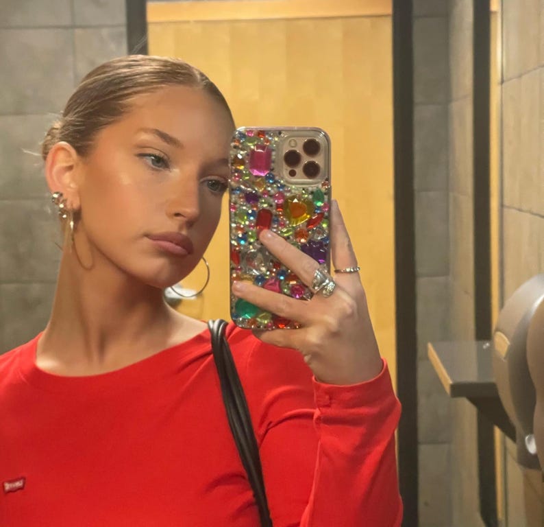May include: A red long-sleeve top. The person is holding a bedazzled phone case with colorful gems. The person is wearing gold hoop earrings and rings. The background is a bathroom with a mirror.