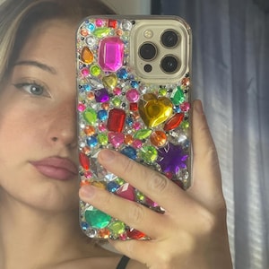May include: A clear phone case bedazzled with an assortment of colorful rhinestones. The gems are various shapes and sizes, including pink, red, yellow, green, and purple. The phone case has a gold camera ring.