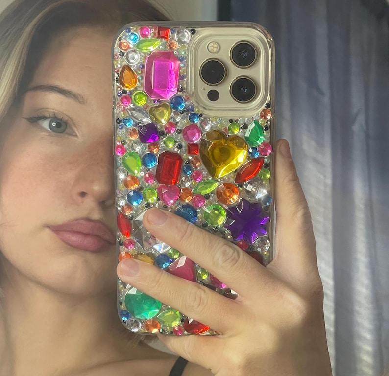 May include: A clear phone case bedazzled with an assortment of colorful rhinestones. The gems are various shapes and sizes, including pink, red, yellow, green, and purple. The phone case has a gold camera ring.