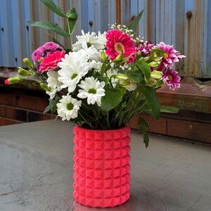 Puede incluir: Un jarrón rosa impreso en 3D con un patrón texturizado contiene un ramo de flores blancas y rosas. Las flores incluyen margaritas blancas, gerberas rosas y claveles rosas.