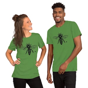 Puede incluir: Dos personas vistiendo camisetas verdes a juego. Cada camiseta presenta una ilustración negra detallada de una abeja en la parte delantera. El diseño de la abeja incluye alas y detalles del cuerpo. Las camisetas son de manga corta.