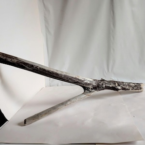 May include: A weathered piece of driftwood, featuring a dark, textured main branch and a lighter-colored, angled secondary branch. The wood displays a mix of dark brown and gray tones, with areas of white. The natural form suggests use in rustic decor.