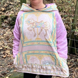 May include: A pink hooded sweatshirt with a colorful carousel horse design on the front. The sweatshirt has a relaxed fit and a large pocket on the front.