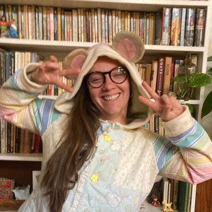 May include: A person wearing a light blue and pink hooded jacket with a mouse ear design. The jacket has a quilted pattern with stars and rainbows. The person is smiling and making a peace sign with both hands.