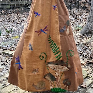 May include: A long brown dress with a corduroy bodice and a patchwork skirt. The skirt features embroidered mushrooms, dragonflies, ferns, and butterflies.